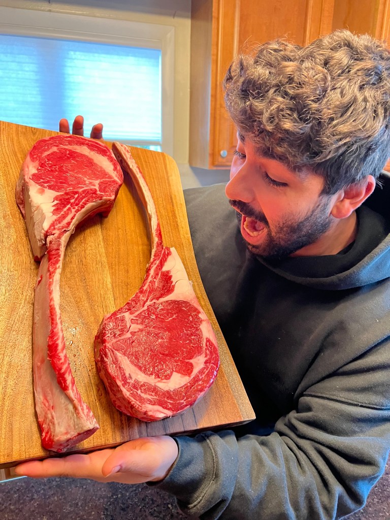 Dom Tuttobene holding two raw tomahawk ribeye steaks on a cutting board